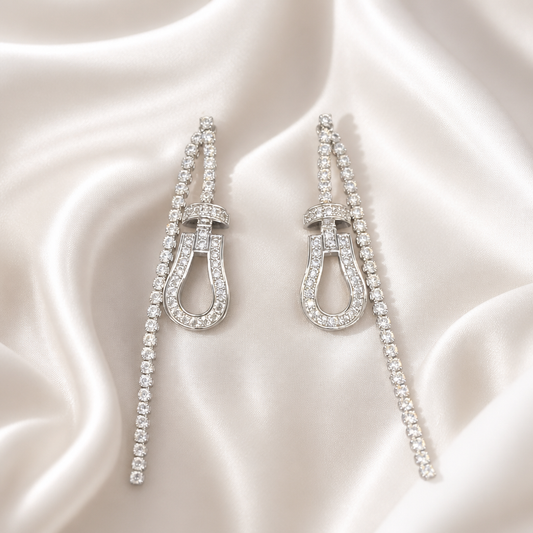 A pair of long drop earrings with horseshoe-shaped centers and rows of clear stones, displayed on draped white satin fabric.