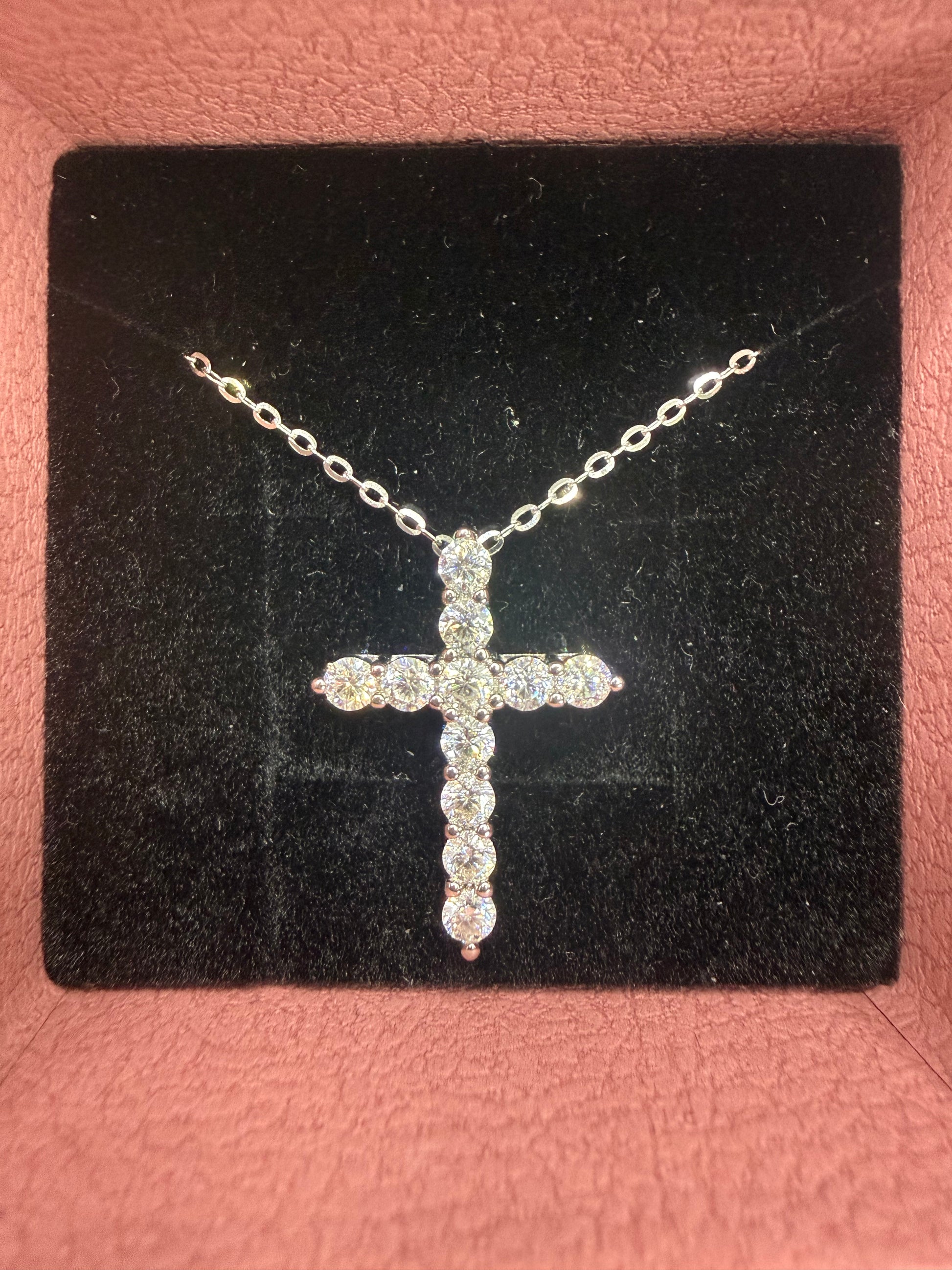 A silver cross pendant necklace paved with clear stones, displayed on a black velvet jewelry box insert against a pink textured background.