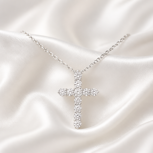 A silver cross pendant necklace paved with clear stones, resting on draped white satin fabric.