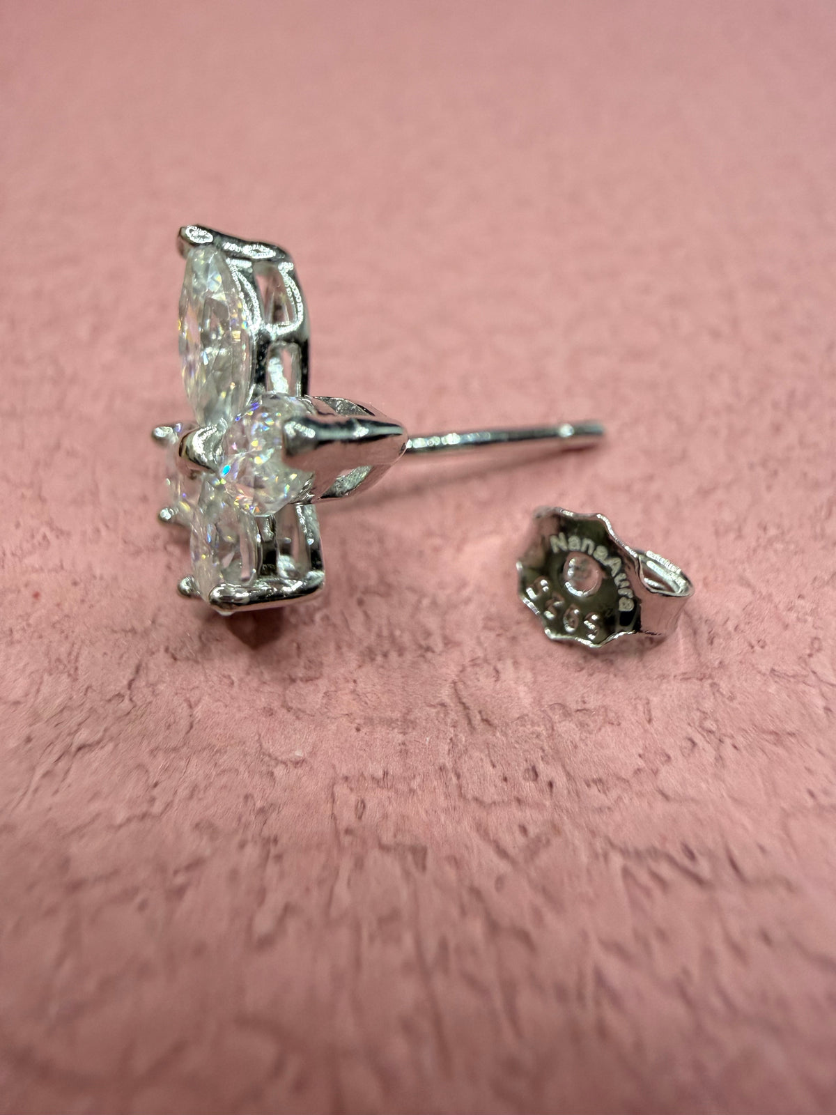 A pair of silver-toned stud earrings with a marquise-cut clear gemstone, shown on a textured pink background. One earring is shown from the front, and the other is shown from the side/back.