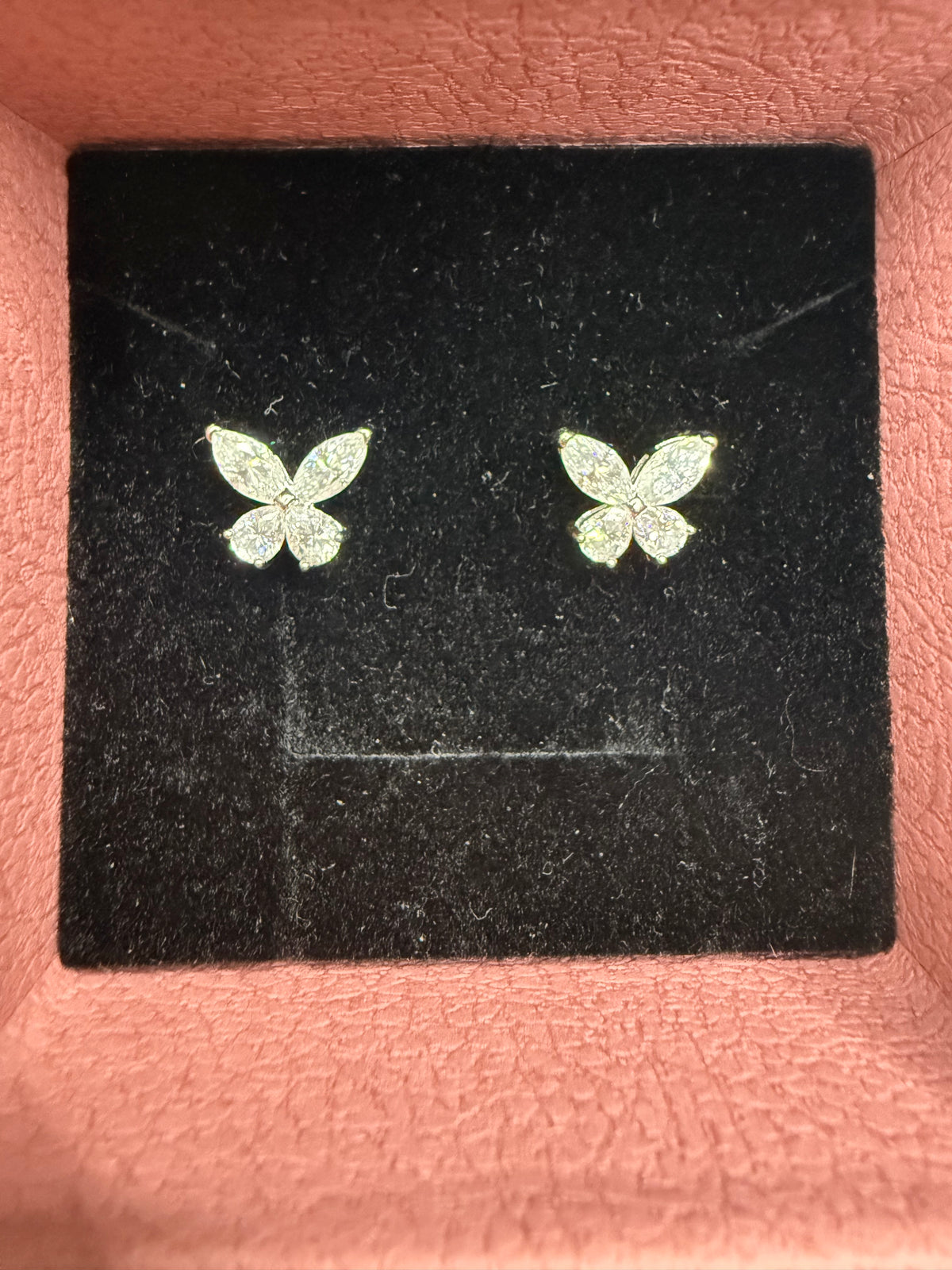 A pair of small butterfly-shaped stud earrings with a textured silver or white glitter finish, displayed on a black velvet jewelry card inside a pink box.
