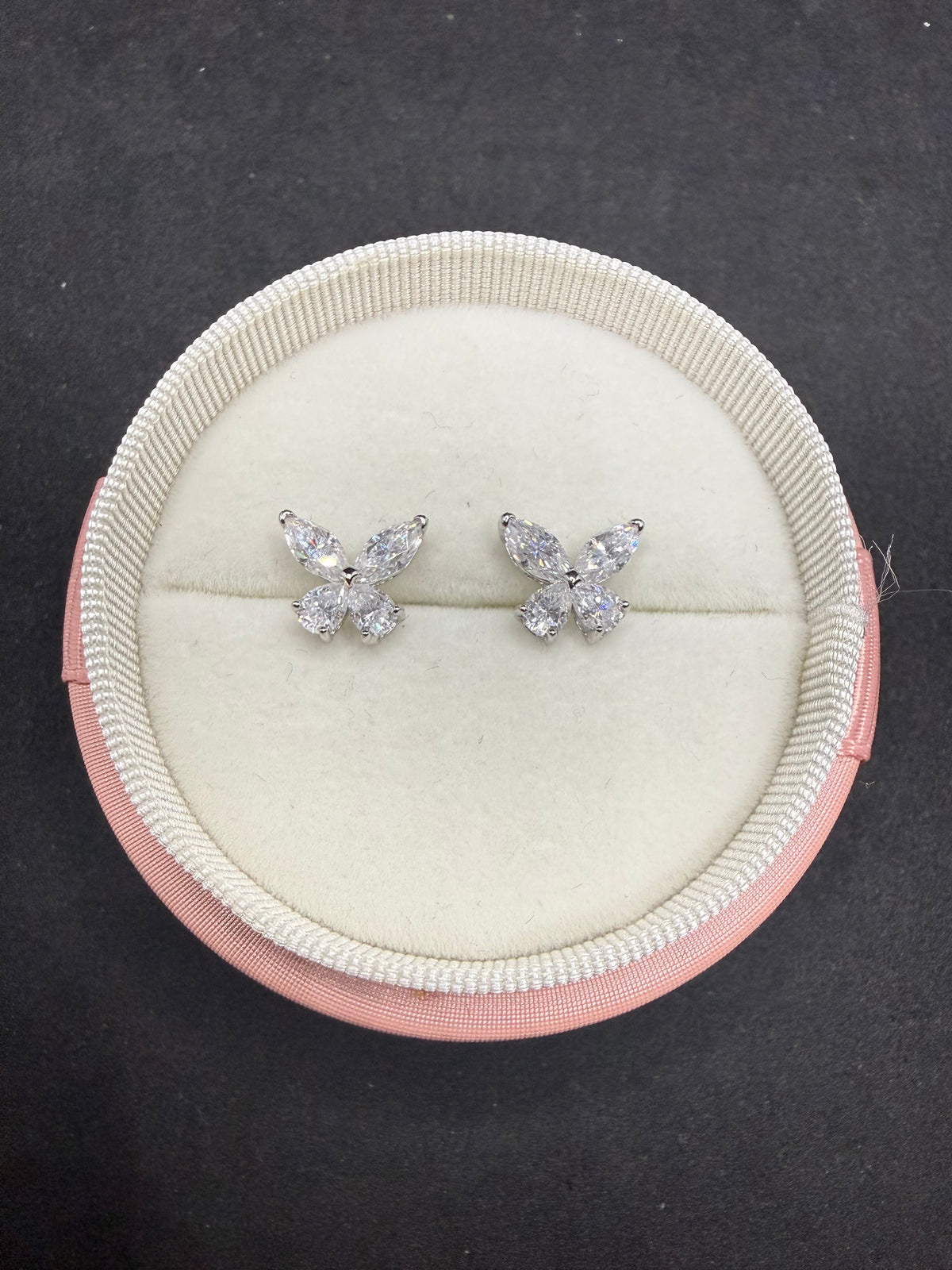 A pair of silver butterfly stud earrings with clear gemstones, presented in a round cream-colored jewelry box with a pink outer rim, set against a dark gray background.