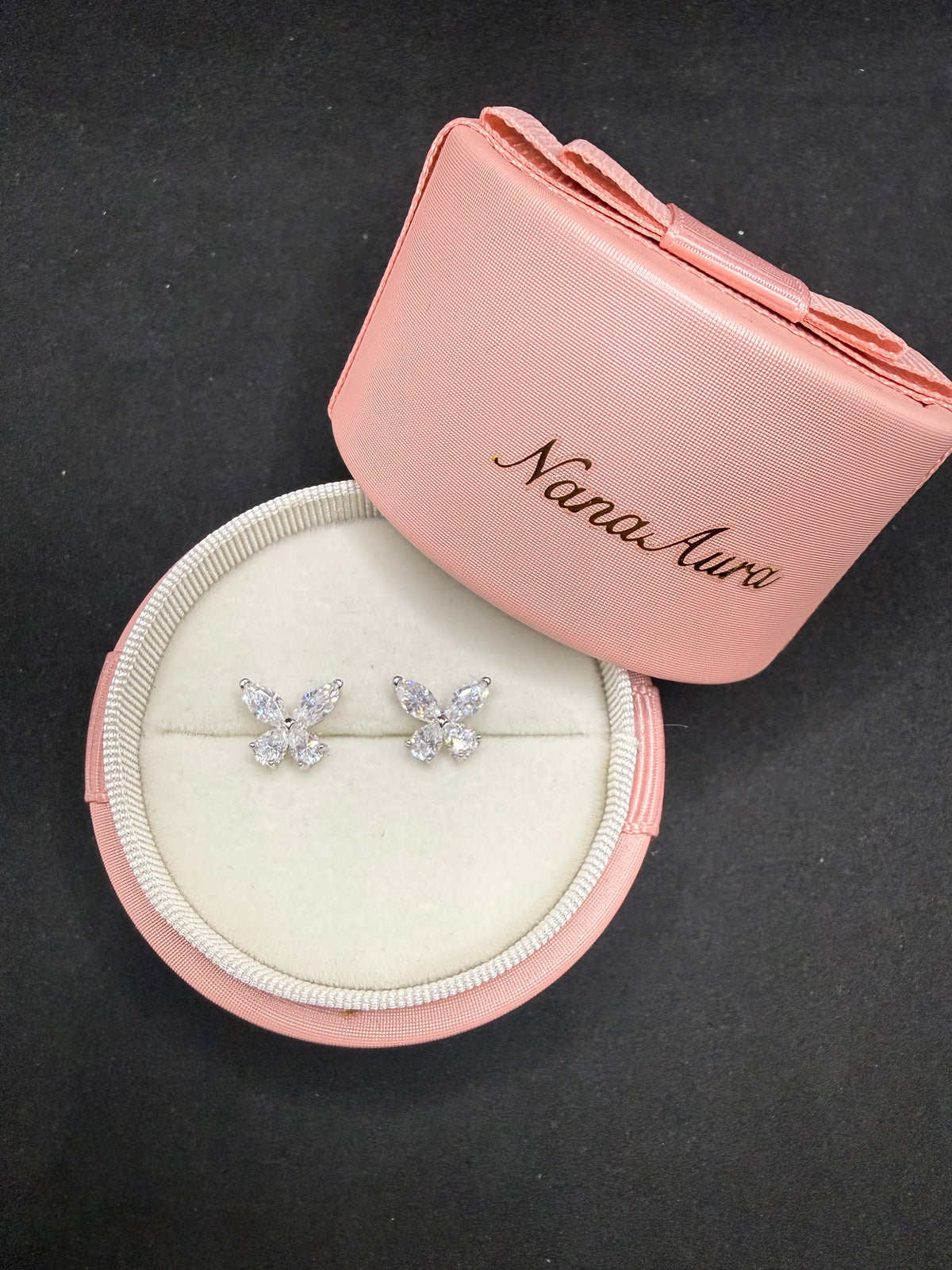 A pair of silver butterfly stud earrings set with clear crystals, presented in an open round pink jewelry box with the brand name 'NanaAuri' printed on the lid, set against a dark background.