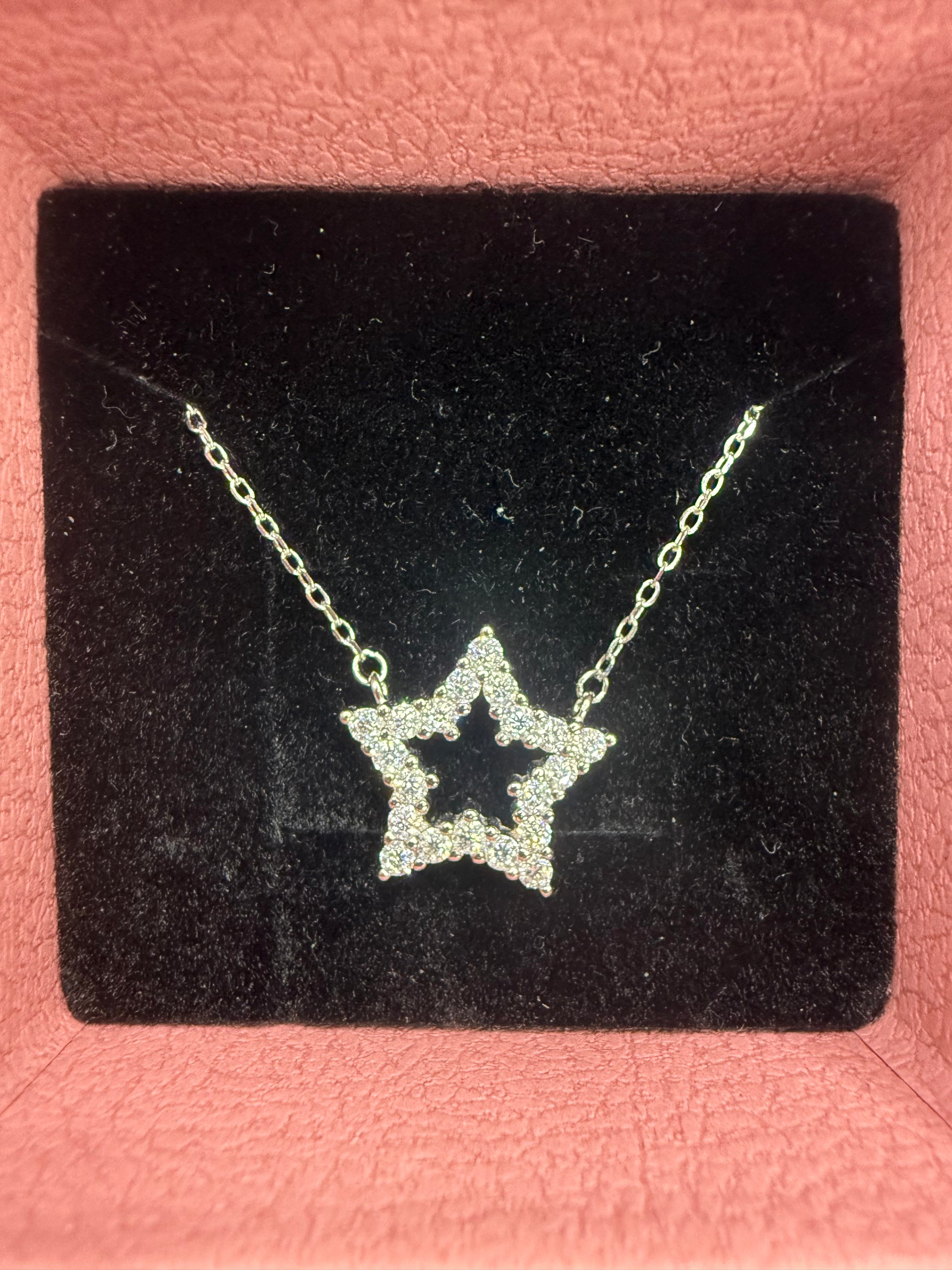 A silver star-shaped pendant necklace with small clear stones, displayed on a black velvet jewelry card against a pink textured background.