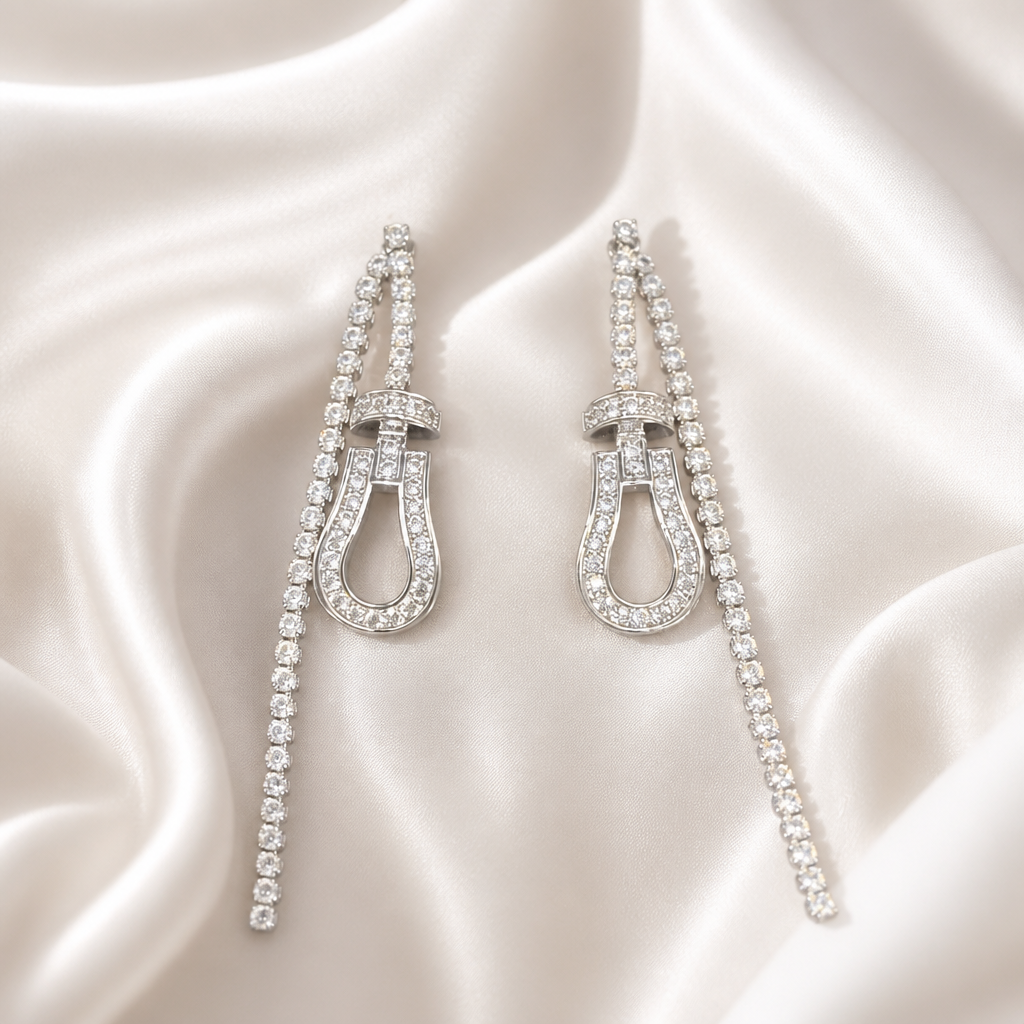 A pair of long drop earrings with horseshoe-shaped centers and rows of clear stones, displayed on draped white satin fabric.