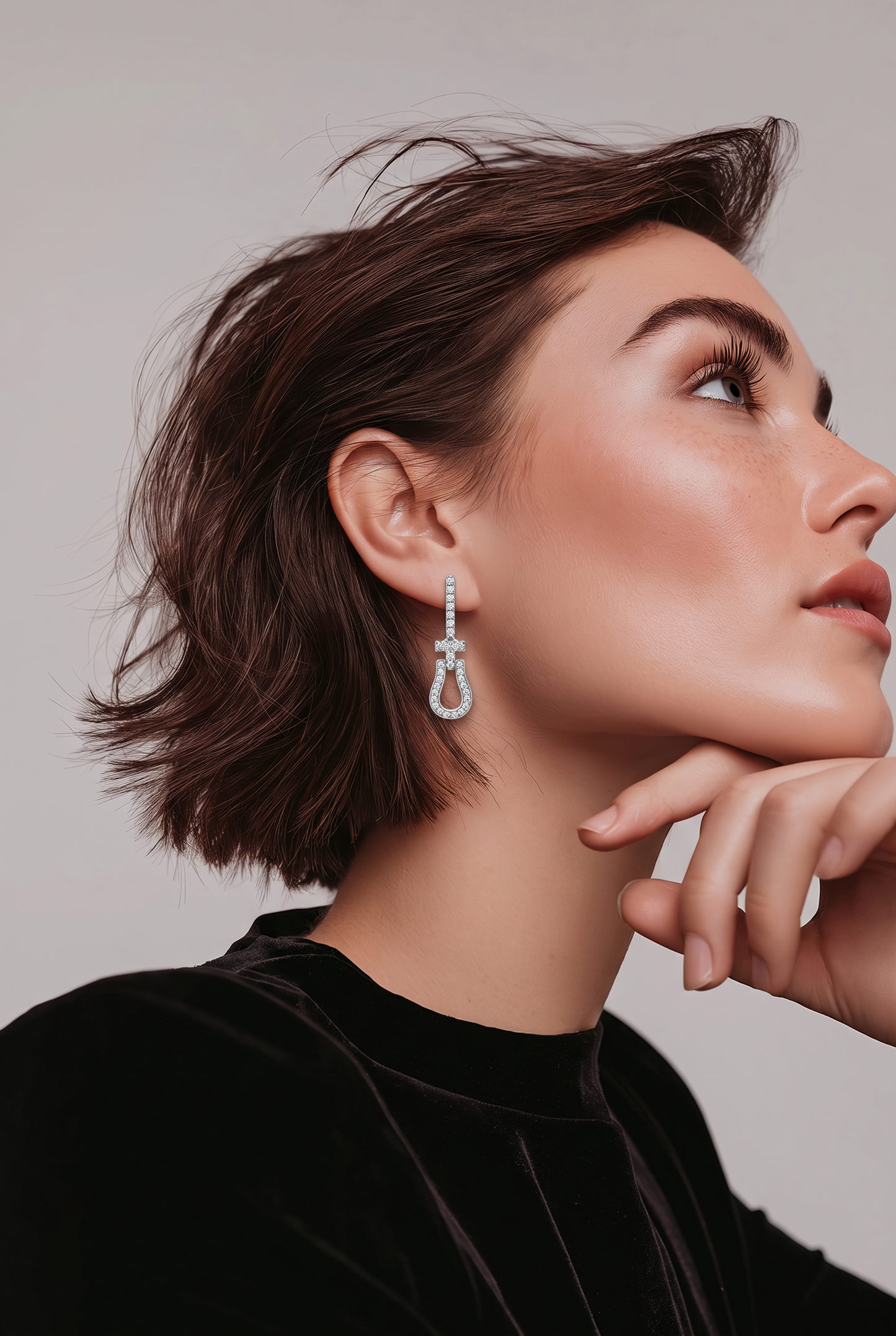 A close-up portrait of a woman with short brown hair wearing a dangling hoop earring with clear stones, against a light gray background.