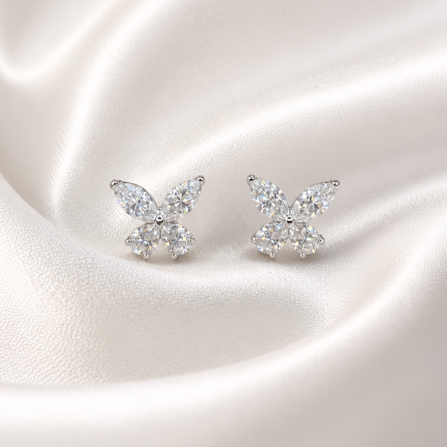 A pair of silver butterfly stud earrings set with clear marquise and round-cut gemstones, displayed on a draped cream satin fabric background.